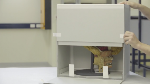 Caption: One side of this box has been left unglued. It is held in place with Velcro. A highly decorated mask made from wool and wood held in a box. One side of the box has been left unglued, with that side secured with Velcro.