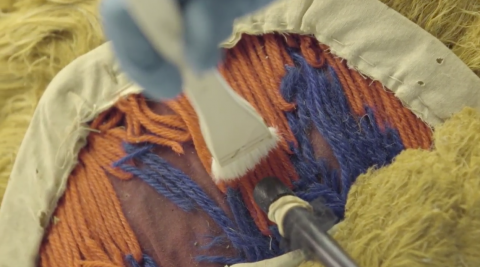 Dust from a mask made from wool is being softly brushed towards the a vacuum cleaner nozzle bounded with netting and a rubber band. 