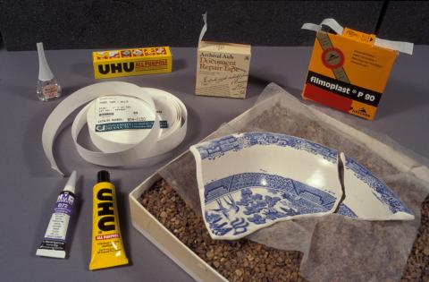A broken ceramic dish surrounded by the materials that will be used to repair it, including glue and adhesive tape.