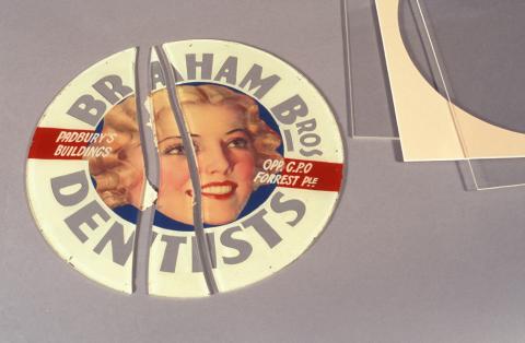 A circular glass advertising sign for 'Braham Brothers Dentists', in the center is an illustration of a smiling woman. The sign has been broken lengthwise into three pieces.